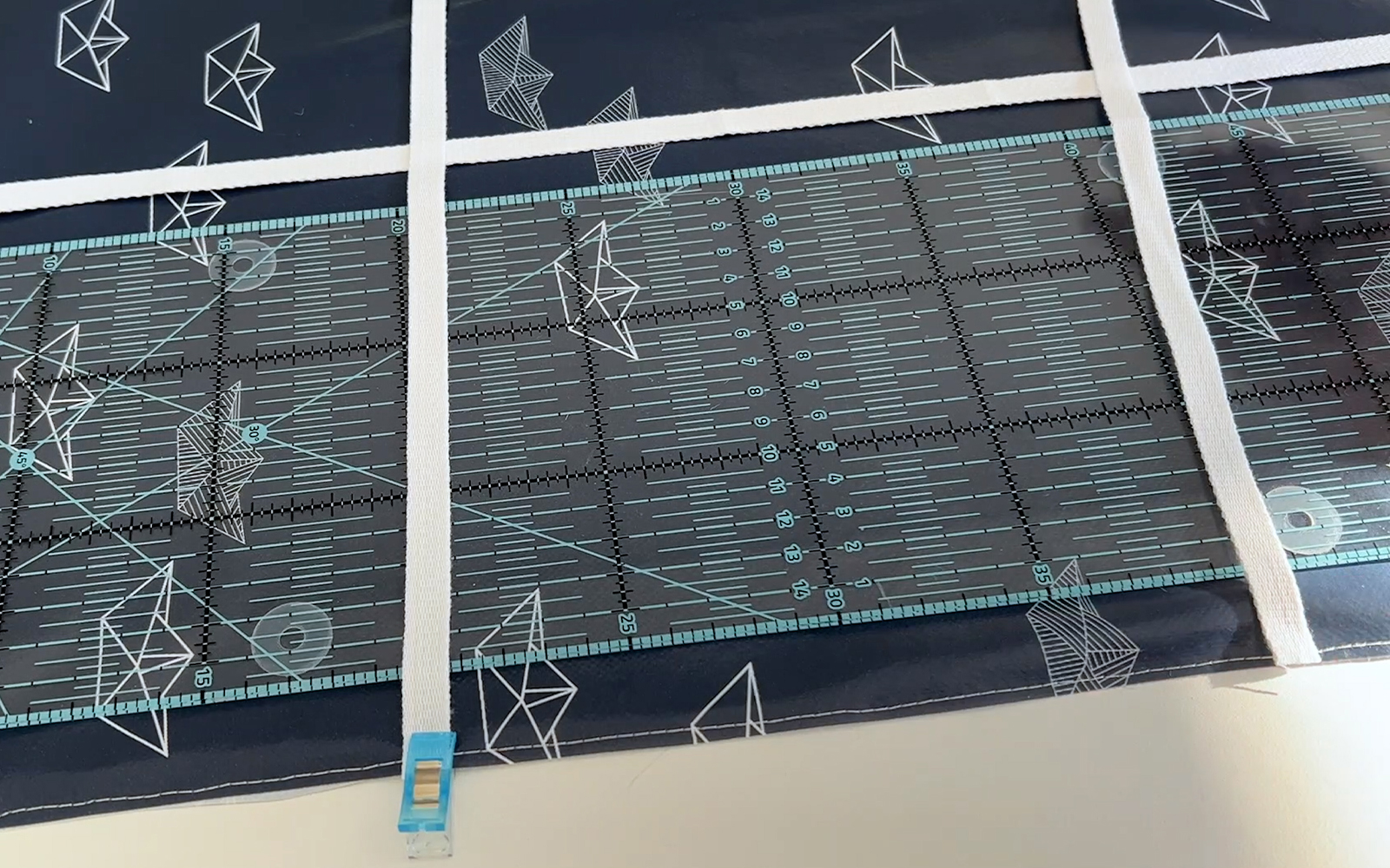 Utilisation de la règle à quilter pour fixer le ruban blanc en lignes droites sur la toile cirée bleu foncé.