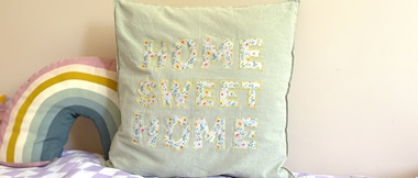 Gros plan sur un coussin brodé « Home Sweet Home » posé contre un mur chaleureux.